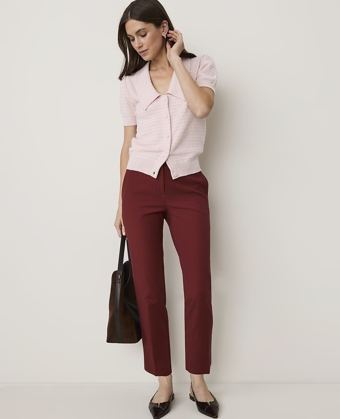A woman in a pink cardigan and maroon pants stands against a plain white background, holding a dark tote bag.