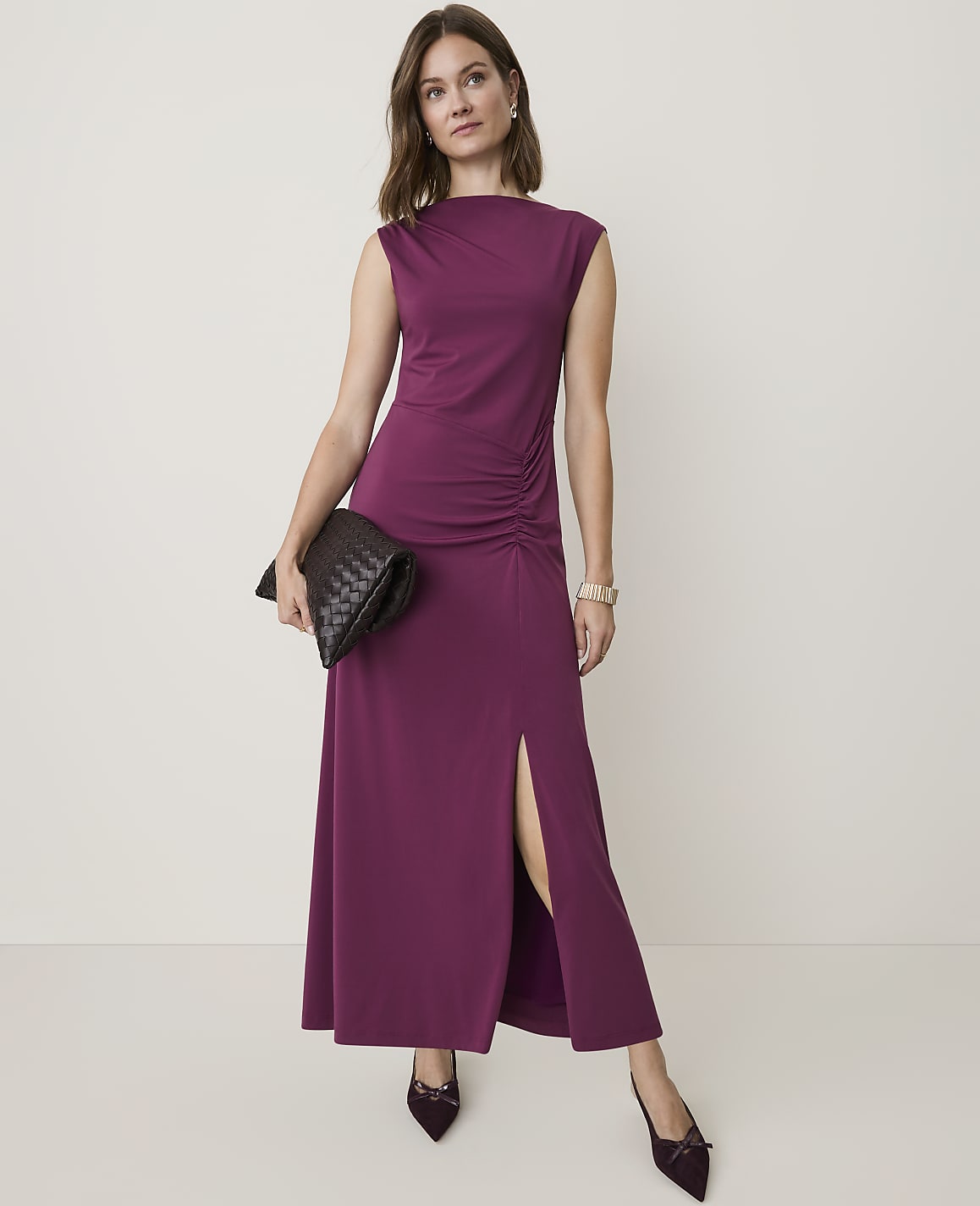 A woman in a plum-colored maxi dress with a high neckline and ruched detailing stands against a plain, light background.