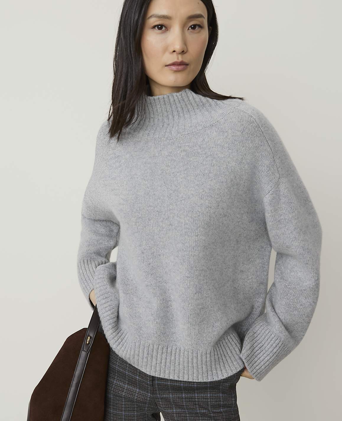 A woman in a grey sweater and plaid pants stands against a plain background, with a brown bag on her left side.