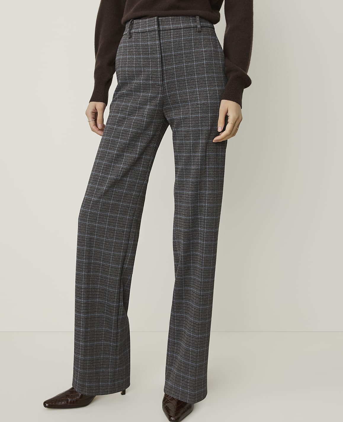 A person stands in a plain, light-colored studio wearing plaid wide-leg trousers and a brown sweater.