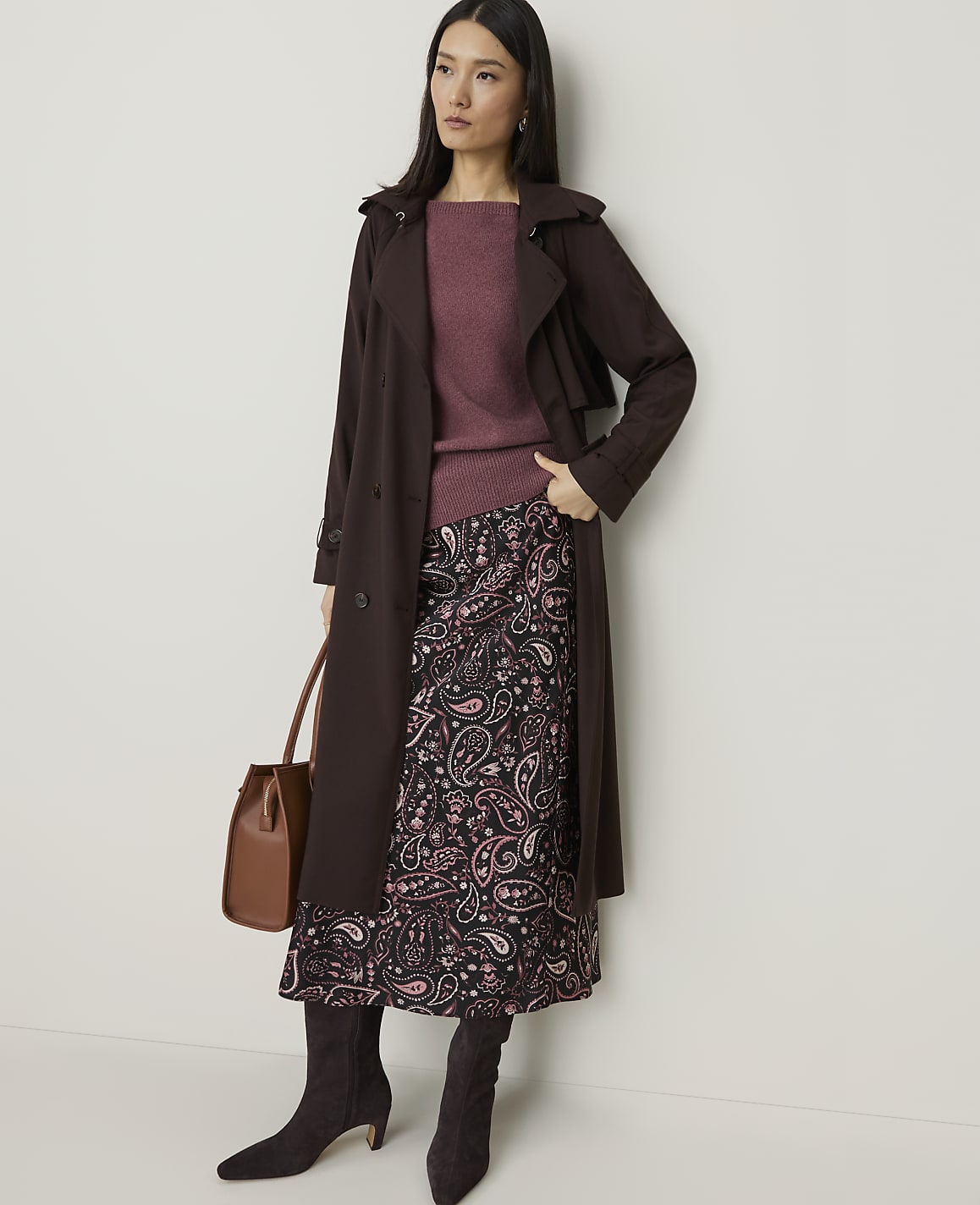 A woman in a brown trench coat, maroon sweater, and patterned skirt stands against a plain white background, holding a brown handbag.