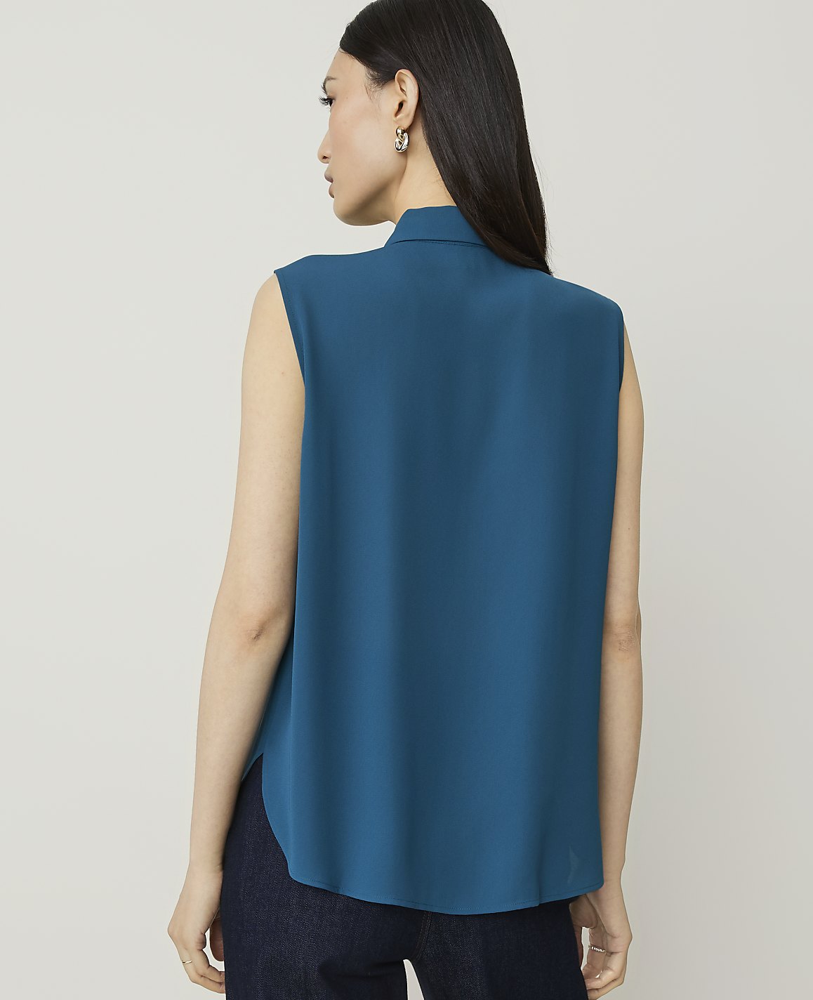A woman in a teal sleeveless top and dark jeans stands against a plain, light gray background.