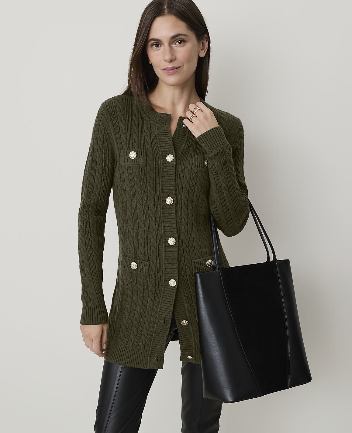 A woman in a brown cable-knit cardigan and black pants stands with a black tote bag in front of a plain, light-colored background.