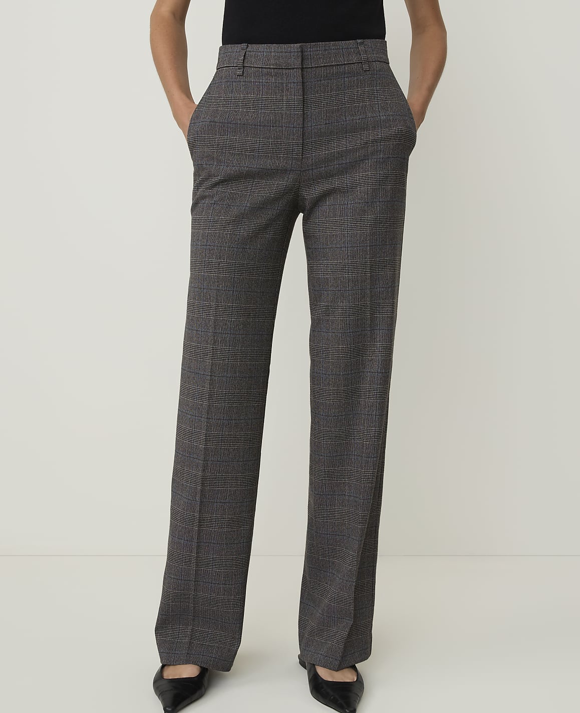 A person stands in front of a plain white background, wearing plaid trousers and black pointed-toe shoes.