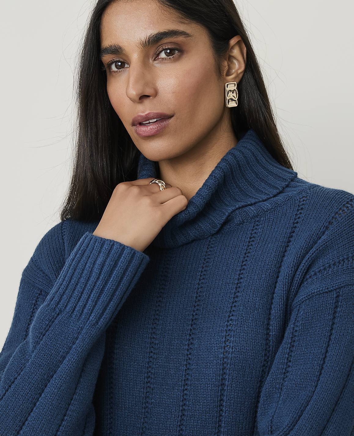 A woman with dark hair and a teal turtleneck sweater is in the foreground, while a plain, light-colored background is visible behind her.