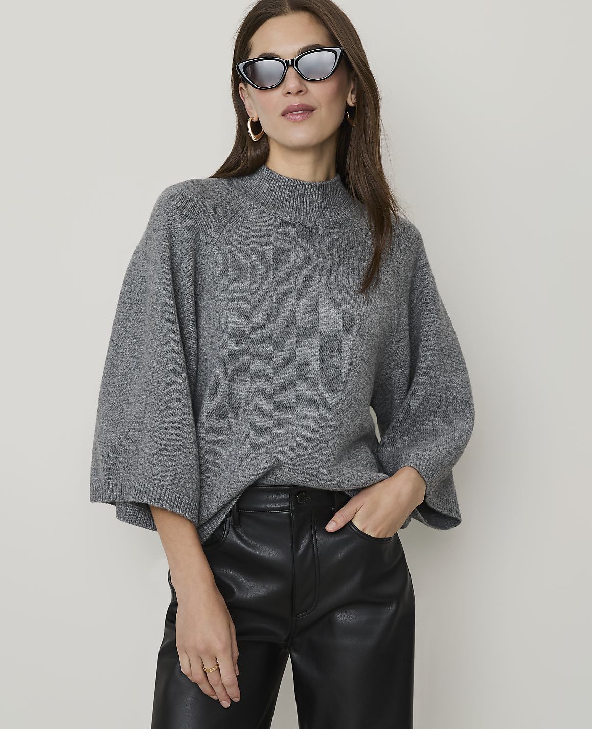 A woman in a grey sweater and black leather pants poses against a plain background.