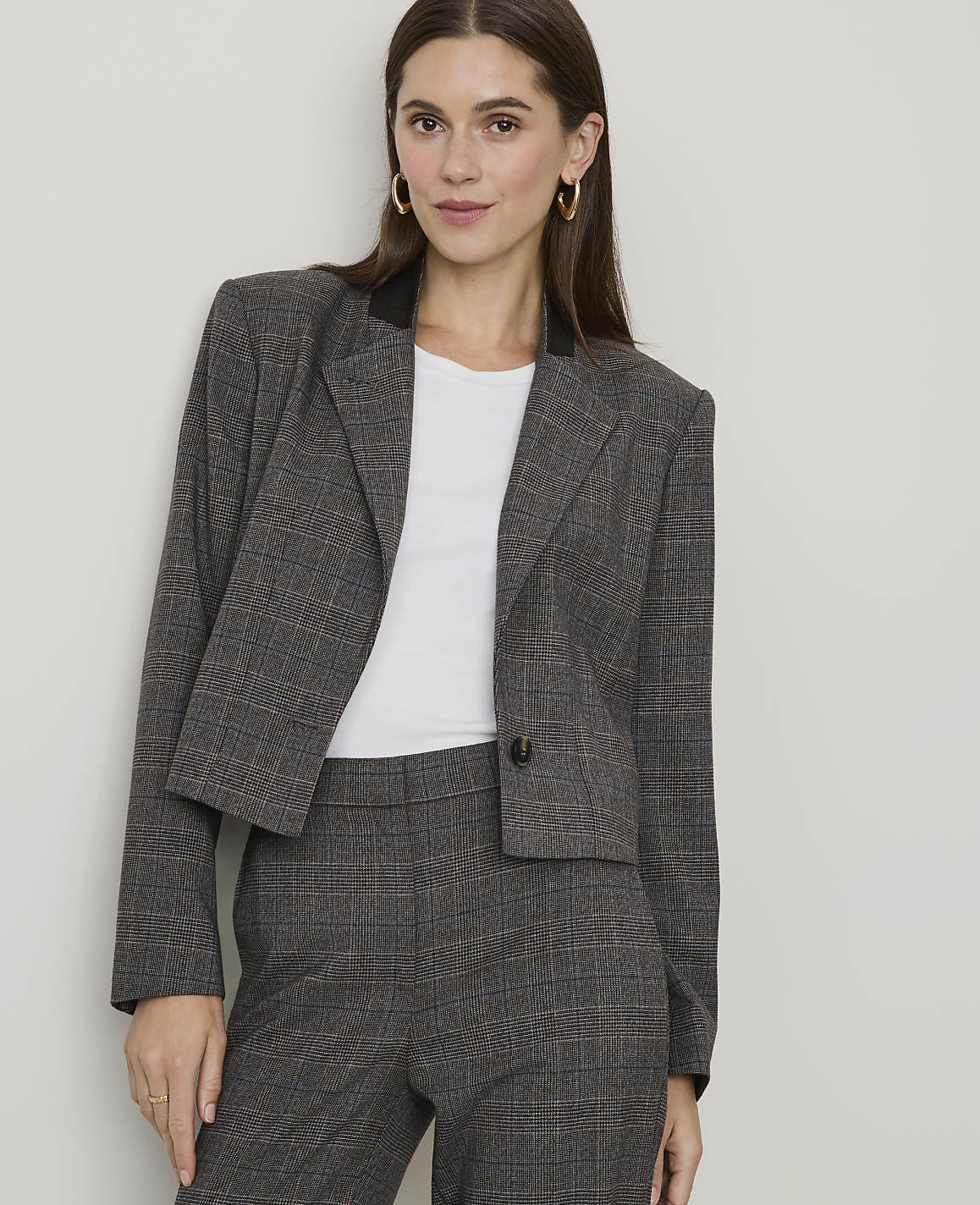 A woman in a plaid blazer and pants stands against a plain, light-colored wall.
