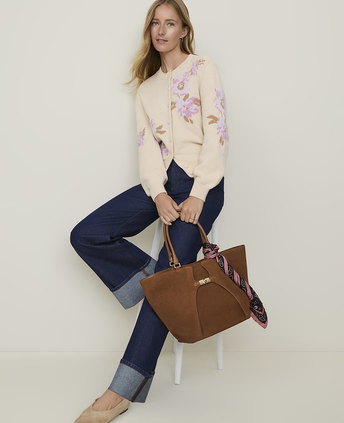 A woman in a cream floral cardigan and dark wash jeans sits on a stool holding a brown suede tote bag, with a plain white background.