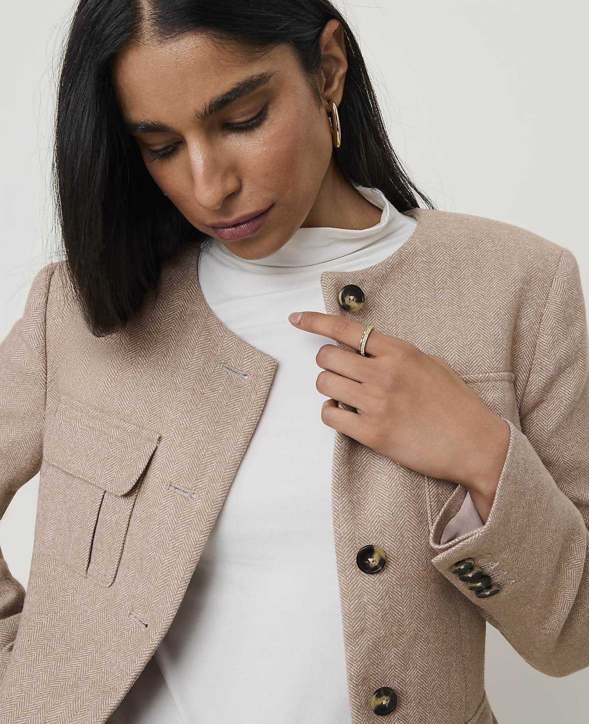 A woman in a beige tweed jacket and white turtleneck stands against a plain, light background.