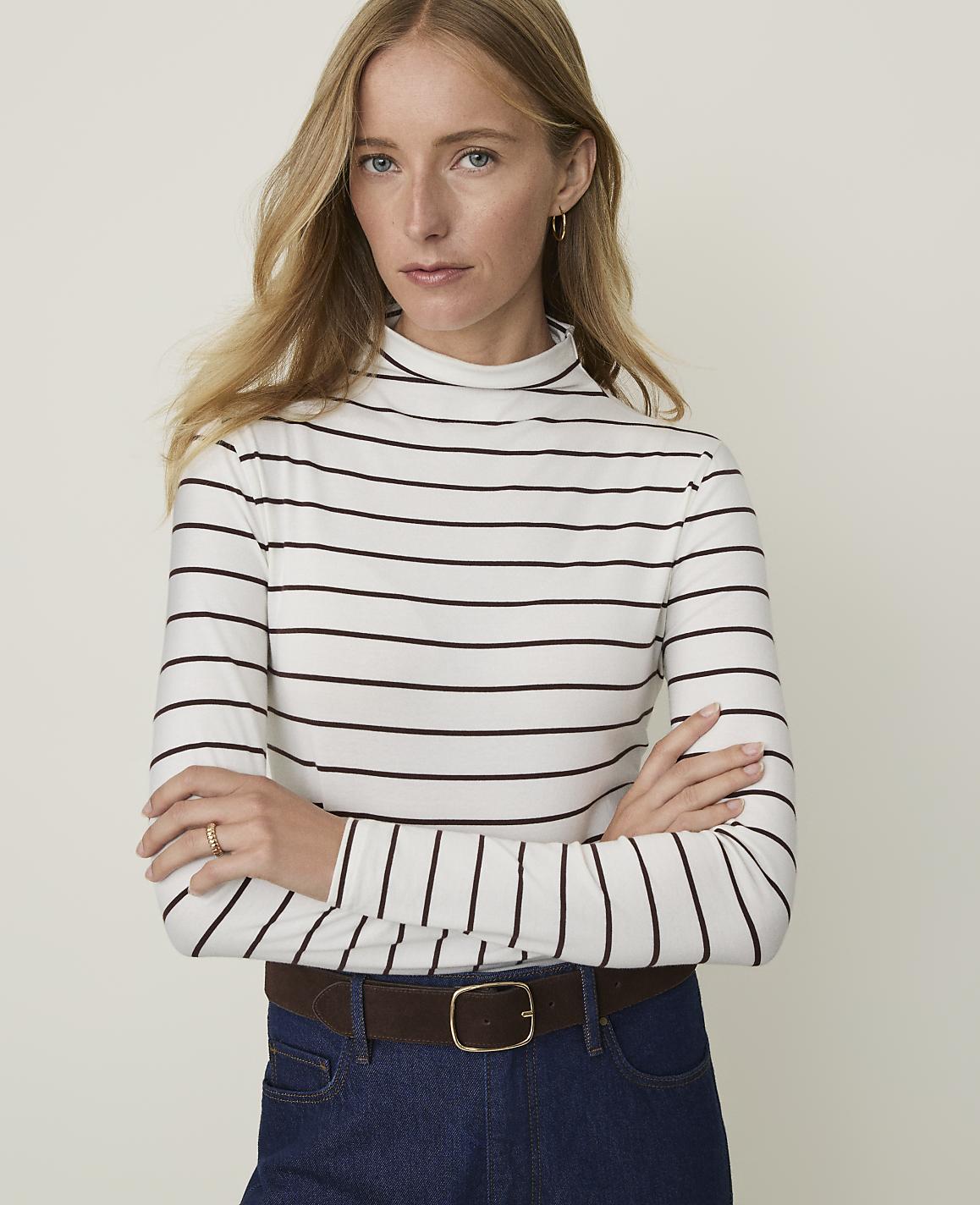A woman with blonde hair and fair skin, wearing a striped turtleneck and dark jeans, stands against a plain, light-colored background.