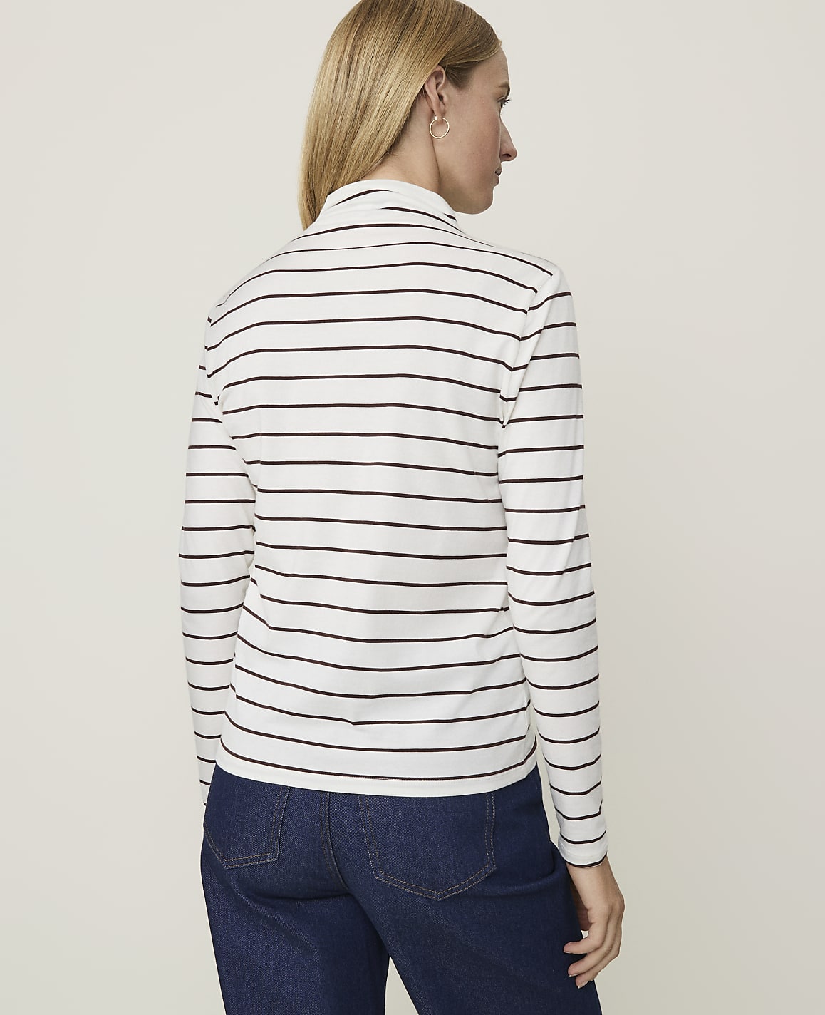 A woman in a striped turtleneck and jeans stands against a plain, light background.