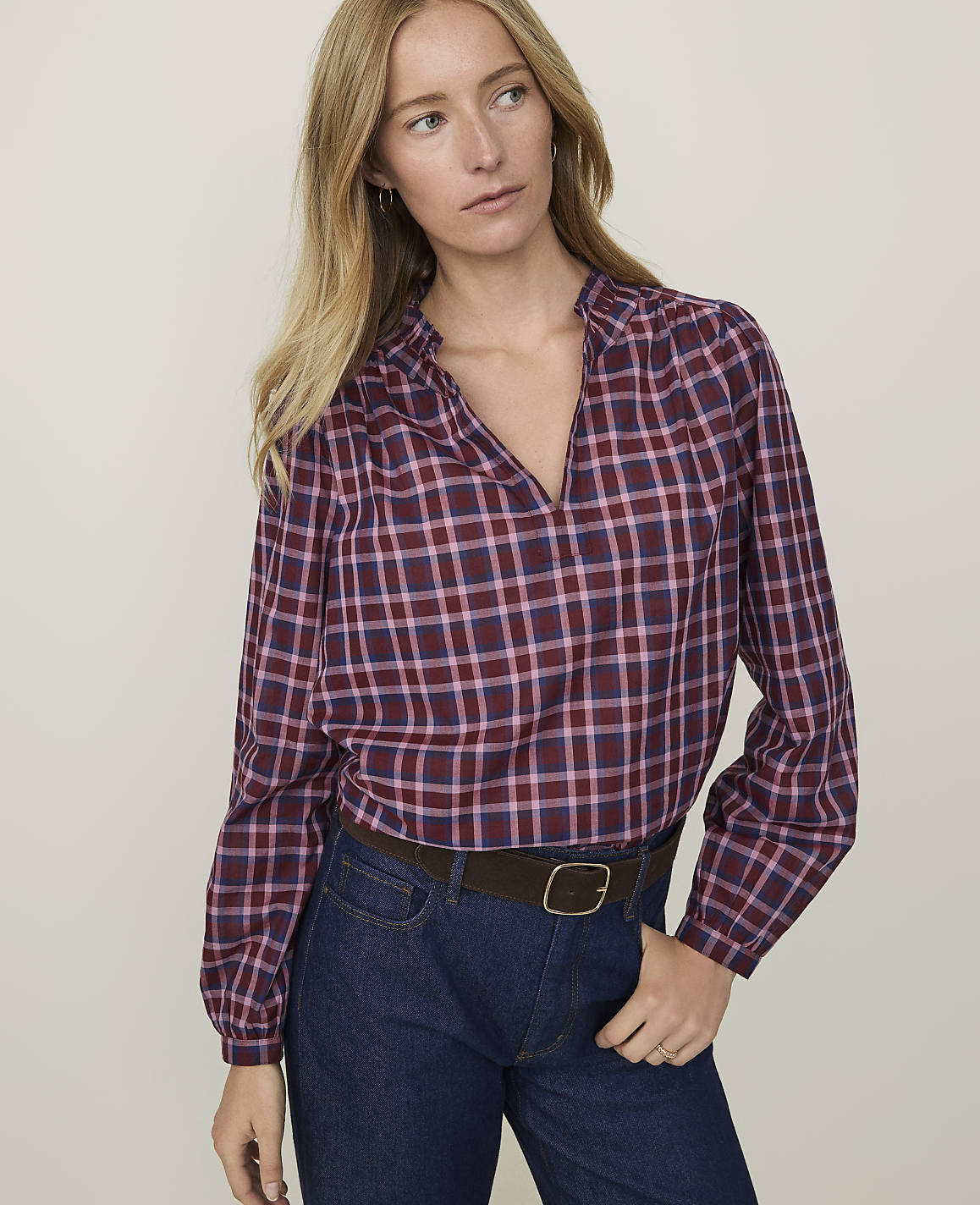 A woman in a maroon and blue plaid blouse and dark jeans stands against a plain, light background.