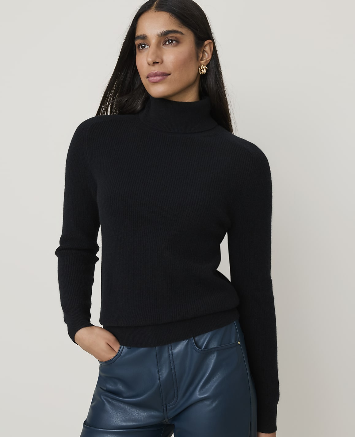 A woman in a black turtleneck and teal pants stands against a plain white background.