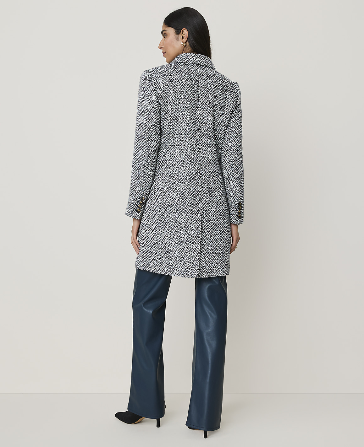 A woman in a patterned coat and teal pants stands against a plain, light-colored background.