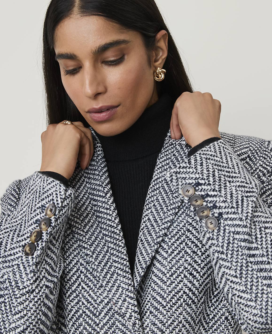A woman in a herringbone blazer and black turtleneck looks down against a plain background.