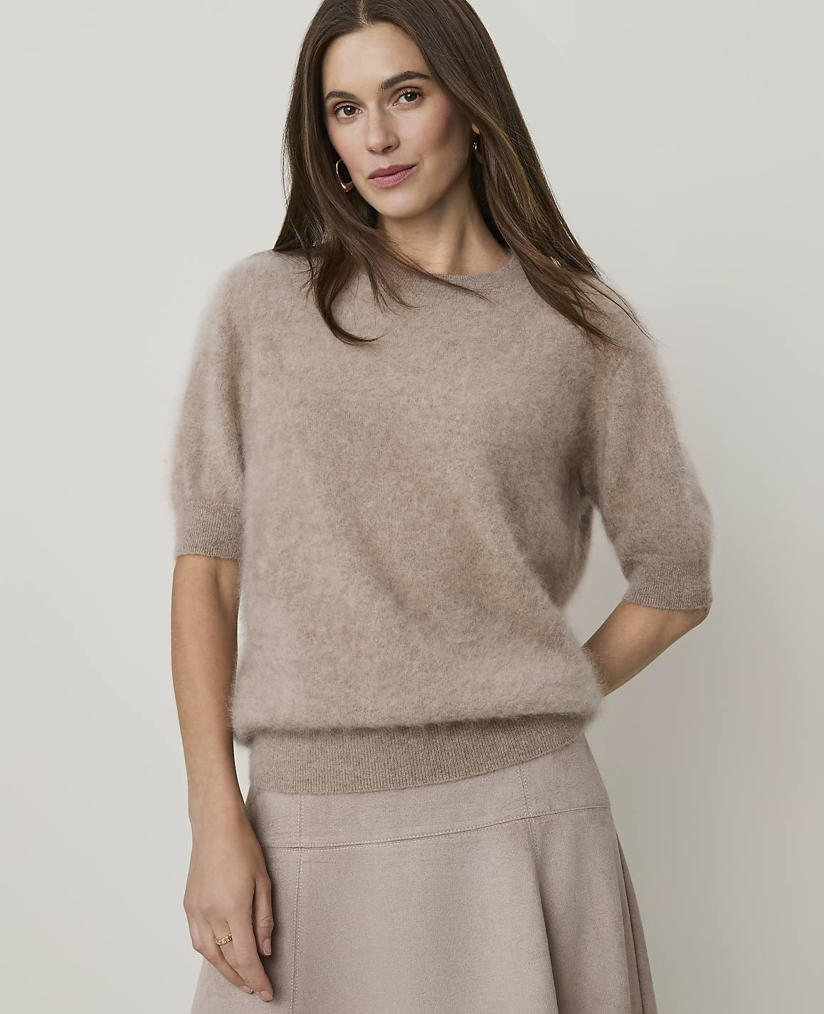 A woman in a fuzzy beige sweater and skirt stands against a plain, light background.