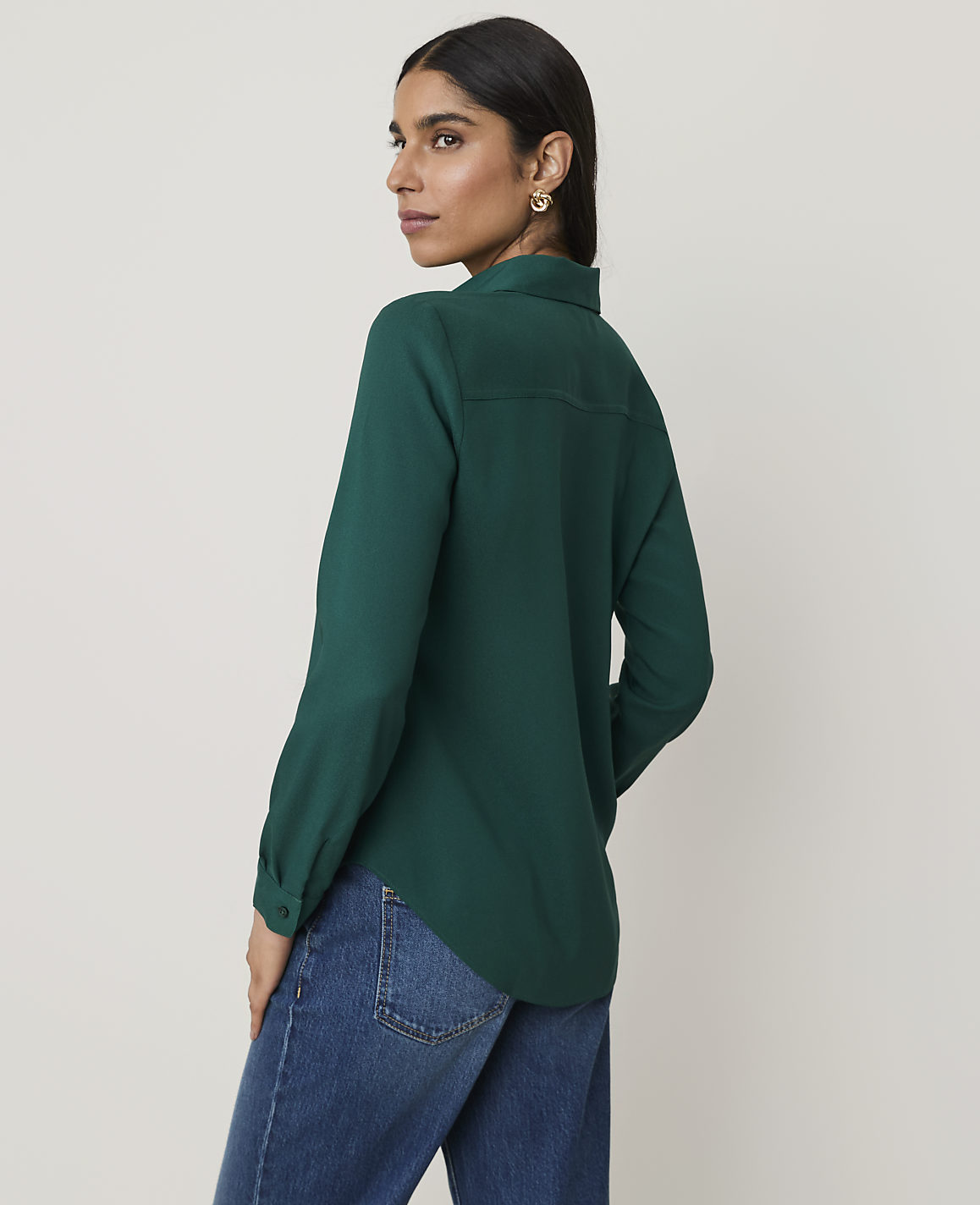 A woman in a green blouse and blue jeans stands against a plain, light-colored background.