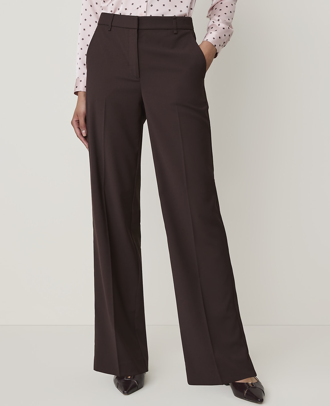 A person in a pink polka dot shirt and brown wide-leg trousers stands against a plain white background.
