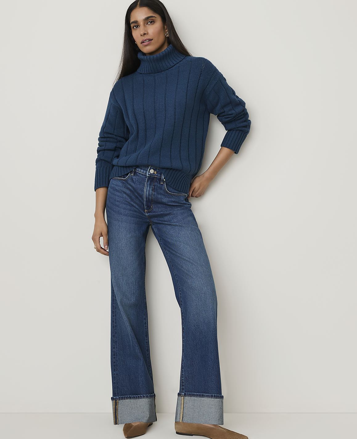 A woman in a teal turtleneck sweater and wide-leg jeans stands against a plain white background.