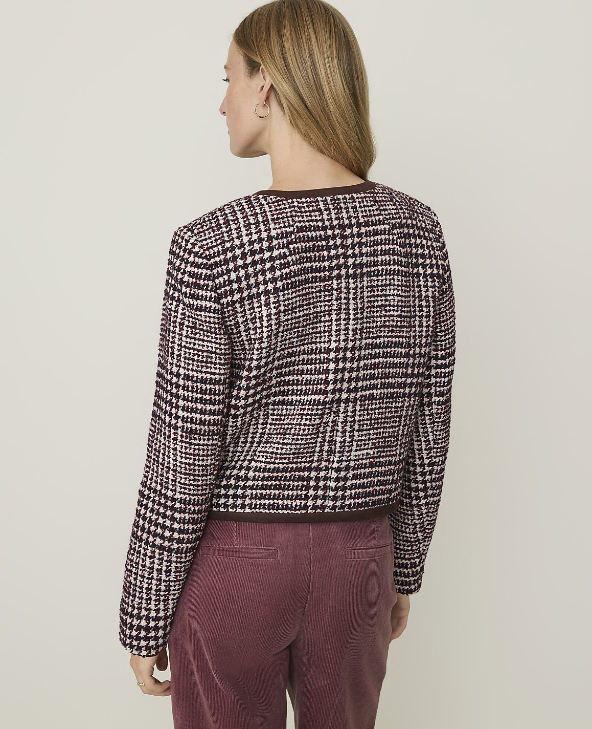 A woman in a plaid jacket and maroon corduroy pants stands against a plain, light-colored background.