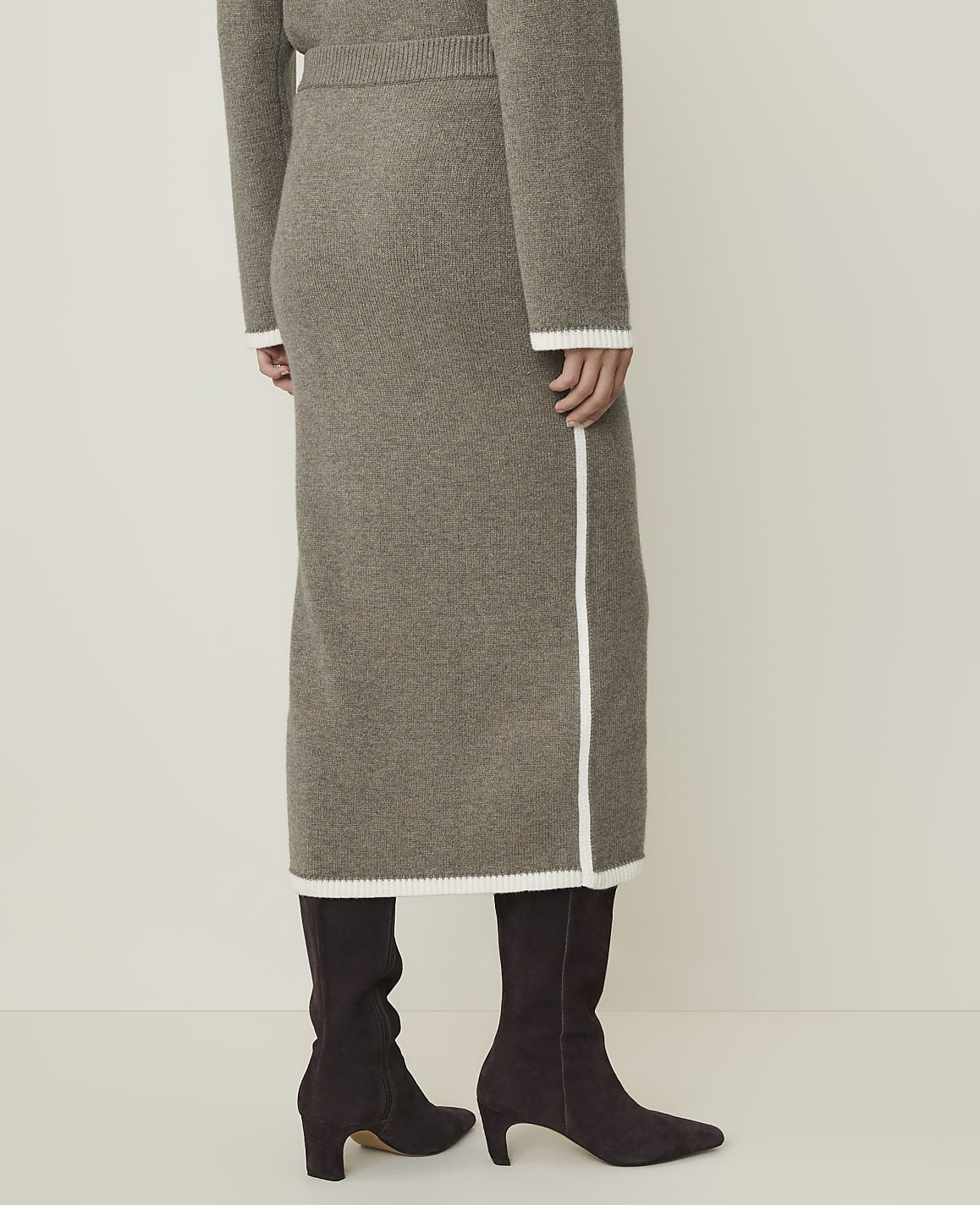A person wearing a taupe knit midi skirt and dark brown boots stands against a plain, light background.