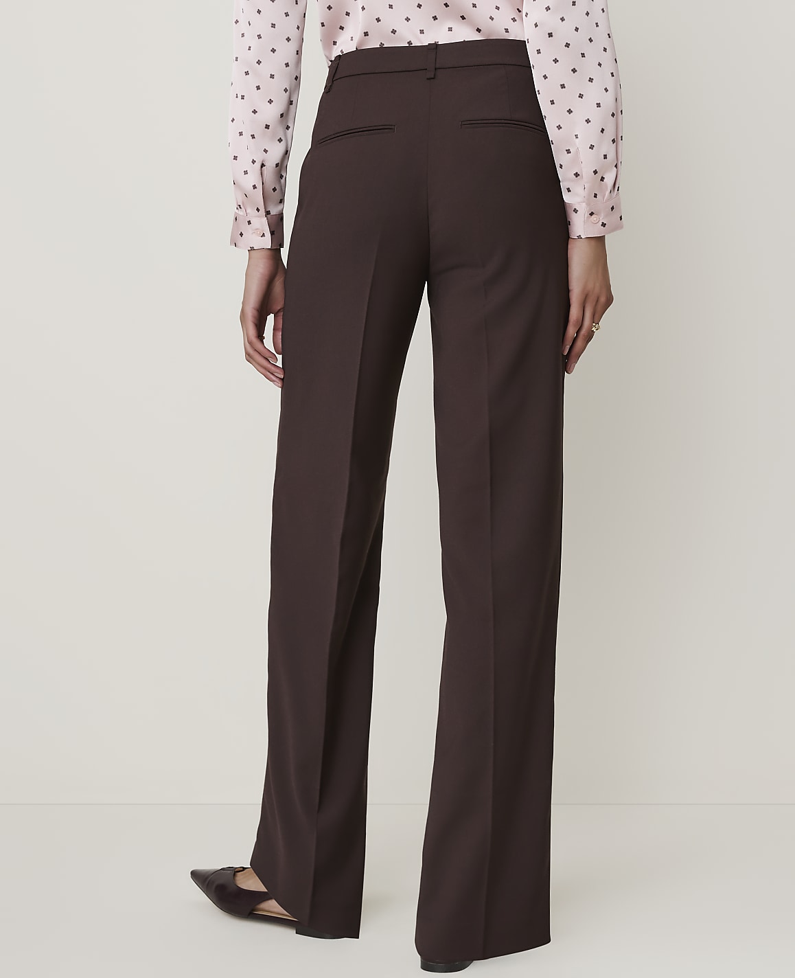 A person wearing brown wide-leg trousers and a pink polka-dot blouse stands against a plain, light-colored background.