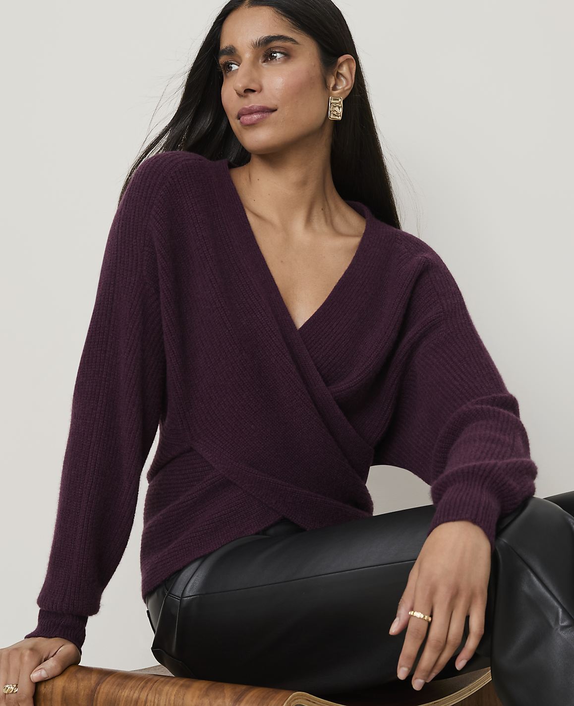 A woman in a plum-colored wrap sweater and black leather pants sits against a plain, light background.