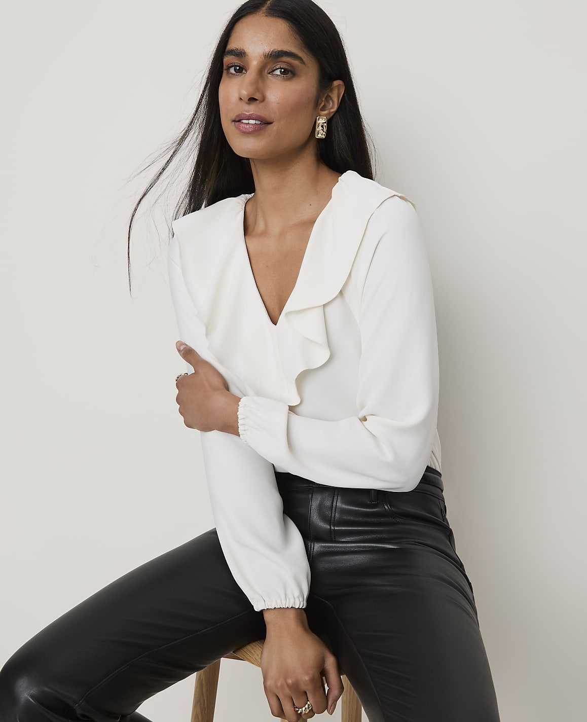A woman in a cream ruffled blouse and black leather pants poses against a plain, light-colored background.