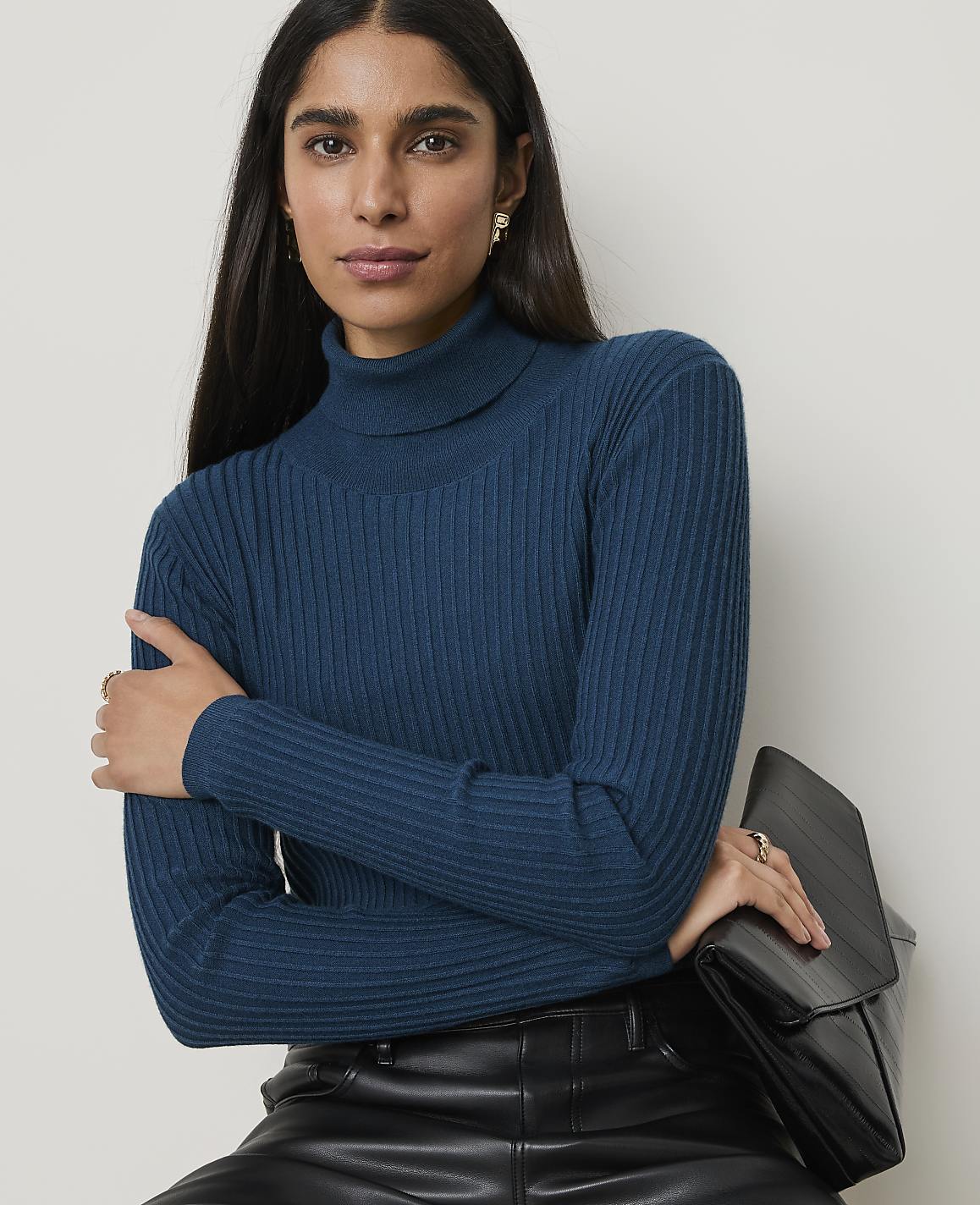 A woman in a teal turtleneck sweater and black leather pants holds a black clutch against a plain white background.