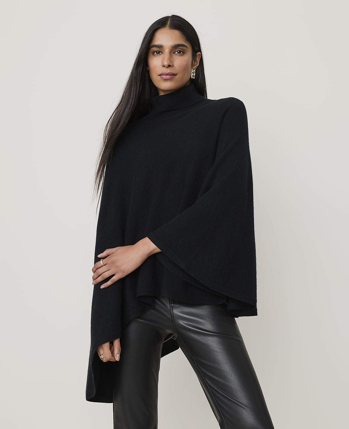 A woman in a black turtleneck poncho and leather pants stands against a plain, light-colored background.