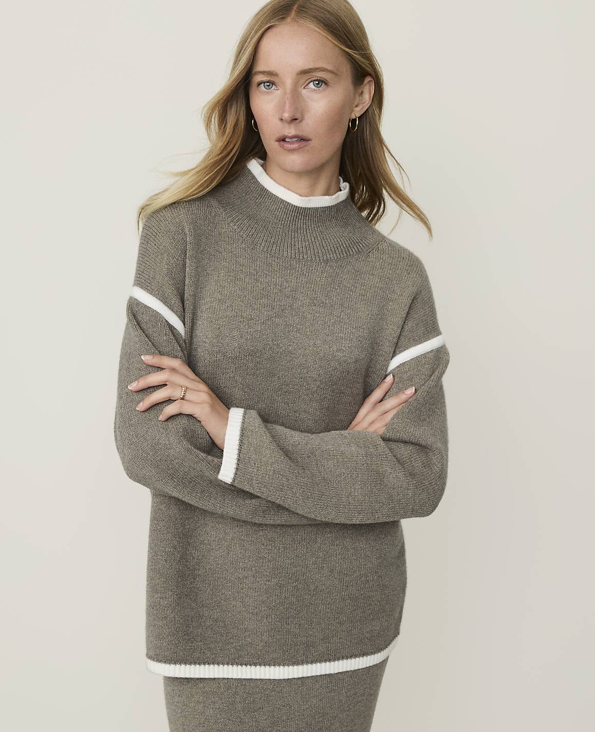 A woman in a taupe sweater with white trim stands against a plain, light-colored background.