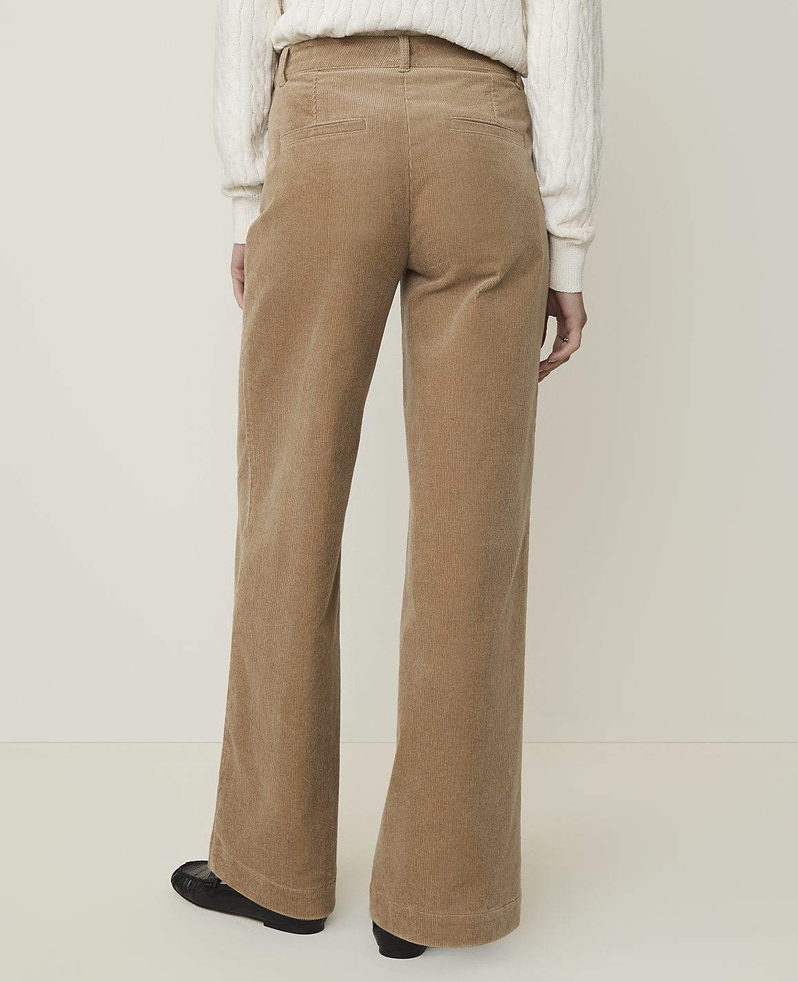 A person wearing tan corduroy pants and a white sweater stands against a plain, light-colored background.