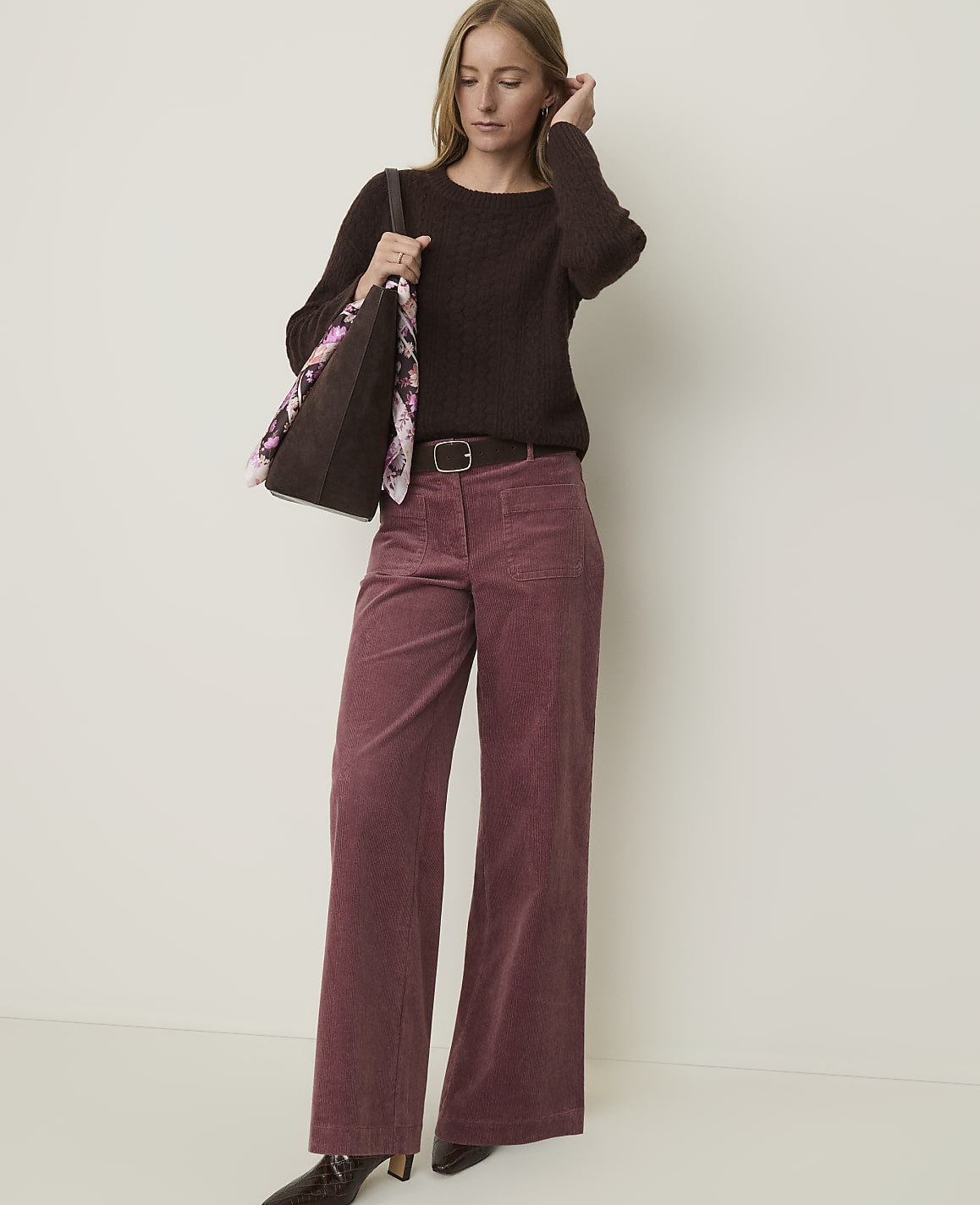 A woman in a brown sweater and pink corduroy pants stands against a plain, light-colored background, holding a tote bag.