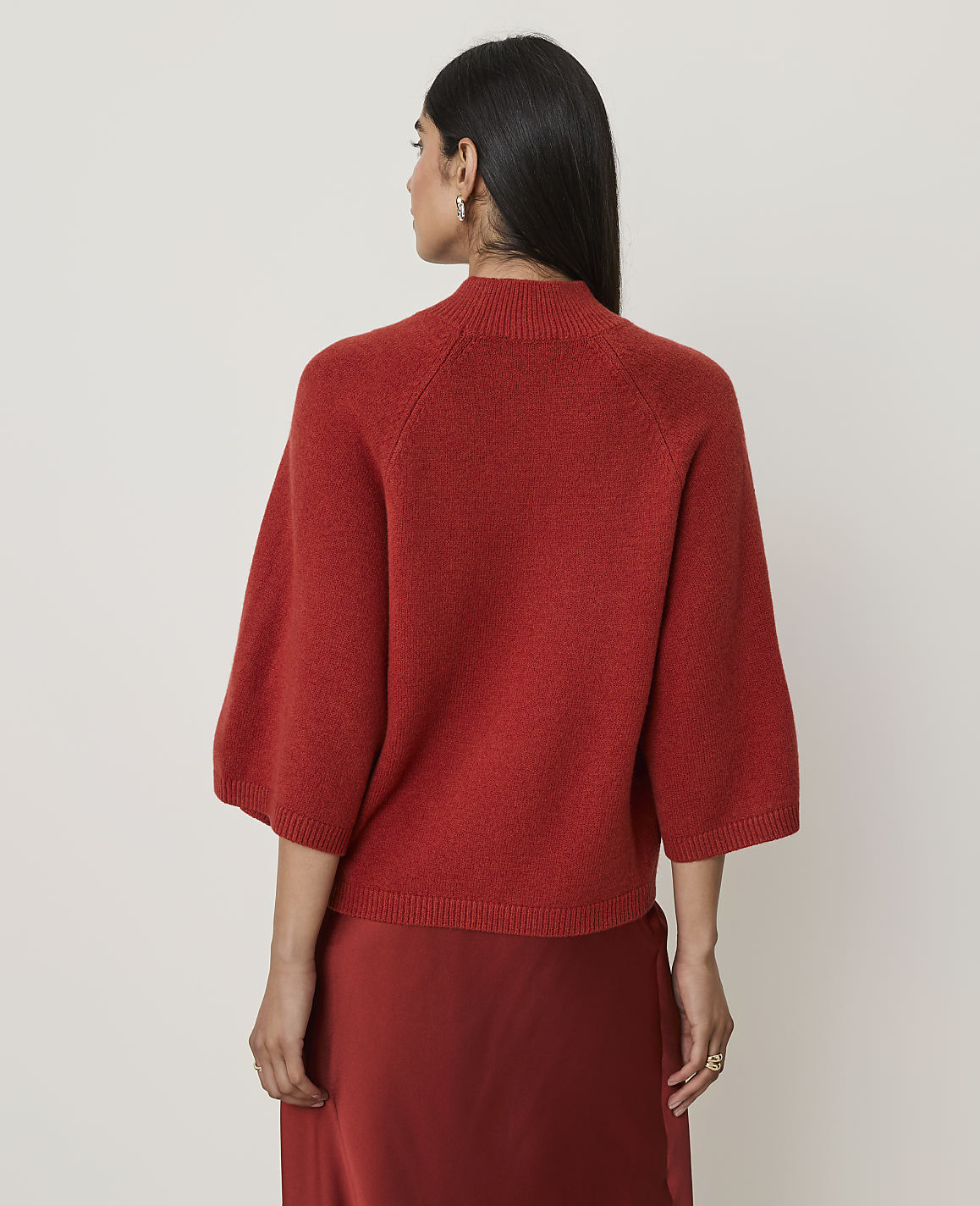 A woman in a rust-colored sweater and skirt stands against a plain white background.