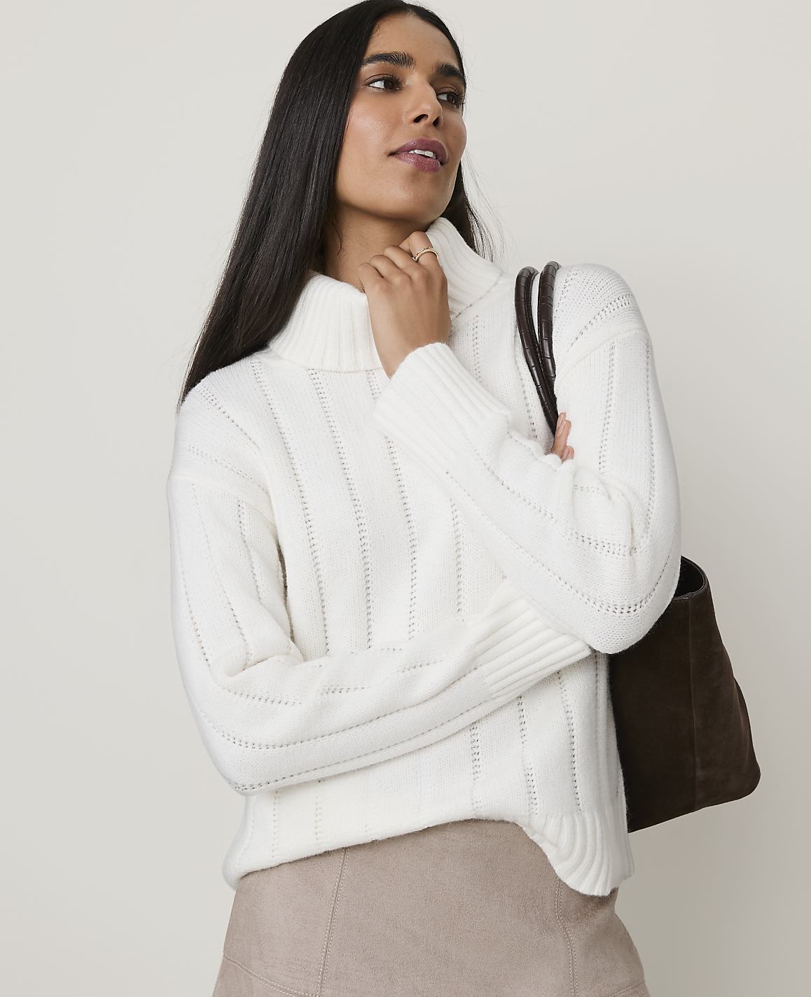 A woman in a cream turtleneck sweater and tan skirt stands against a plain, light background, carrying a dark brown tote bag.