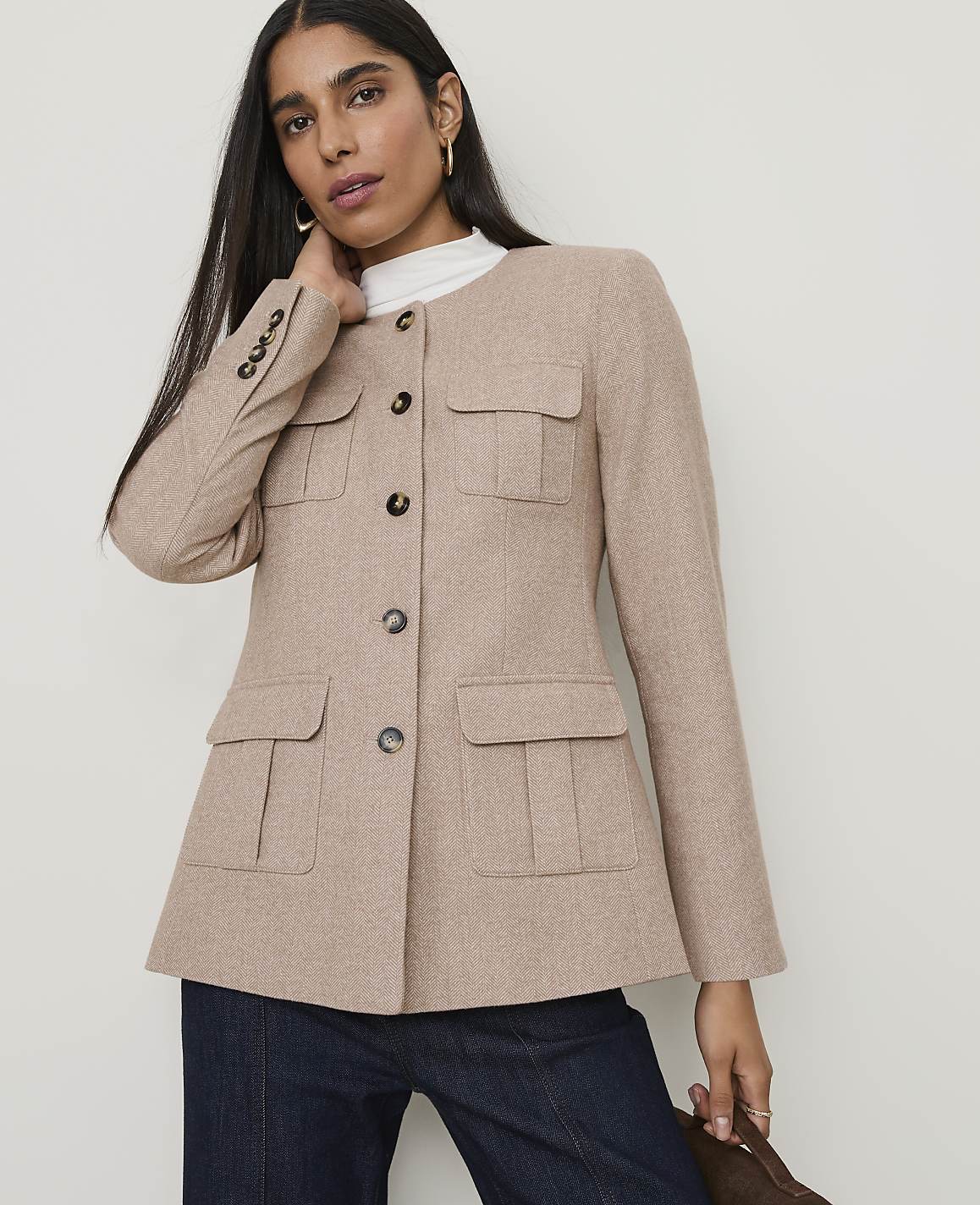 A woman in a tan jacket and dark jeans stands against a plain background.