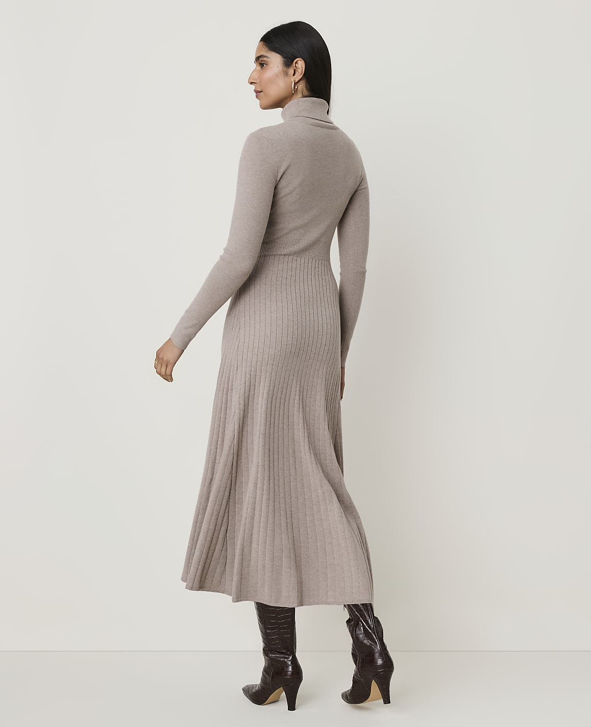 A woman in a beige turtleneck dress and brown boots stands against a plain, light-colored background.