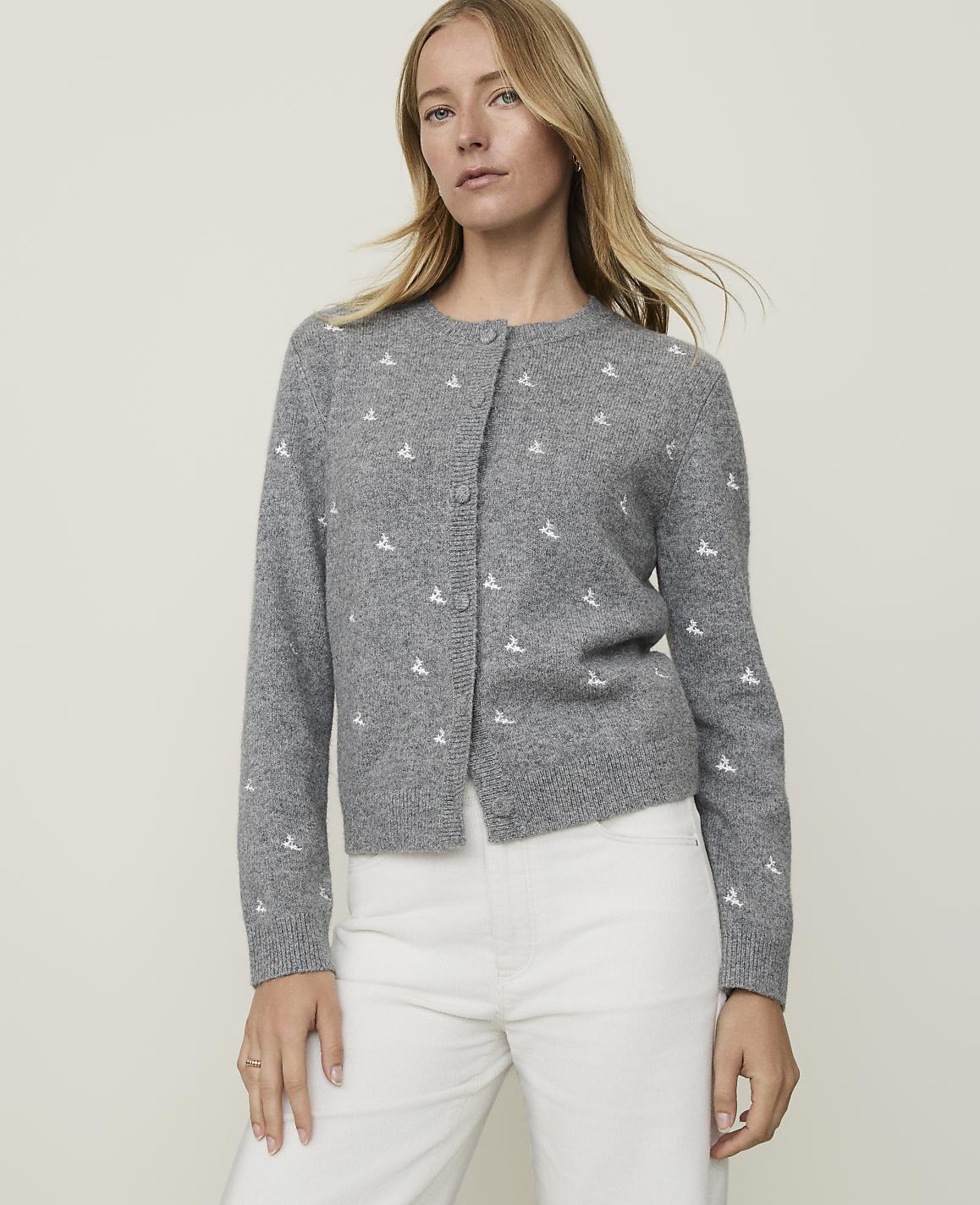 A woman in a grey cardigan with a white embroidered pattern stands against a plain grey background.