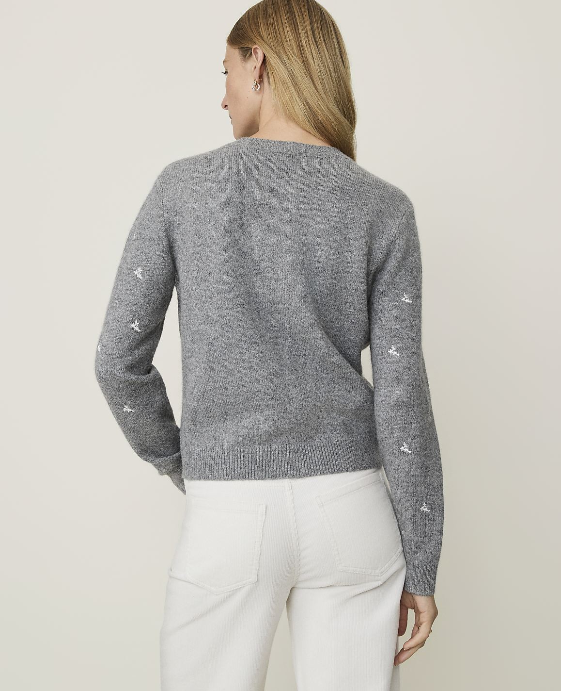 A woman in a grey sweater with embroidered details on the sleeves and white pants stands against a plain, light grey background.