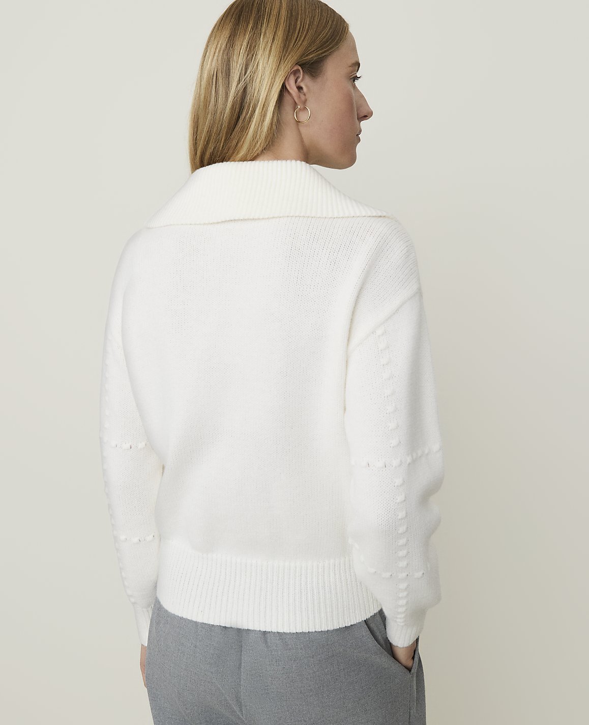 A woman in a white sweater and gray pants stands against a plain, light background.