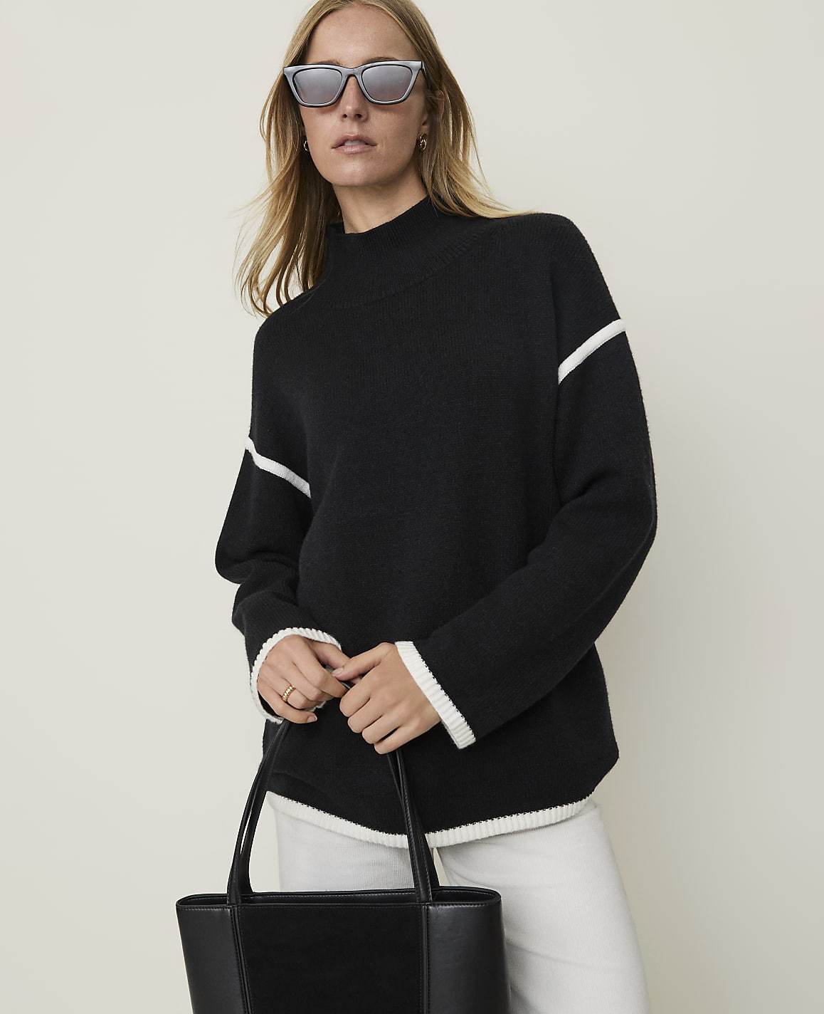 A woman in a black sweater with white trim and sunglasses holds a black tote bag in front of a plain background.