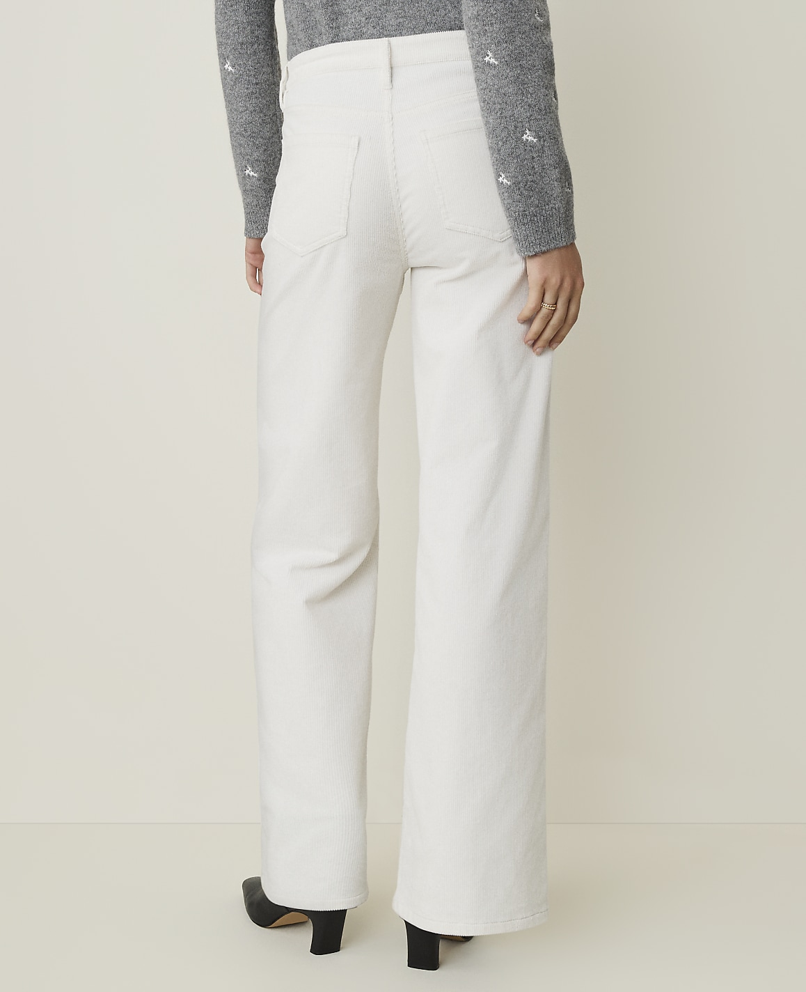 A person wearing white wide-leg pants and a grey sweater stands against a plain, light-colored background.