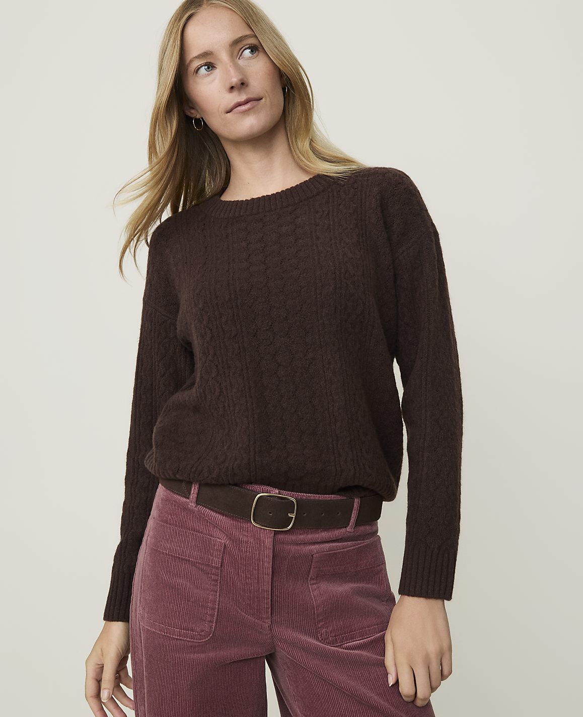A woman in a brown cable-knit sweater and pink corduroy pants stands against a plain white background.
