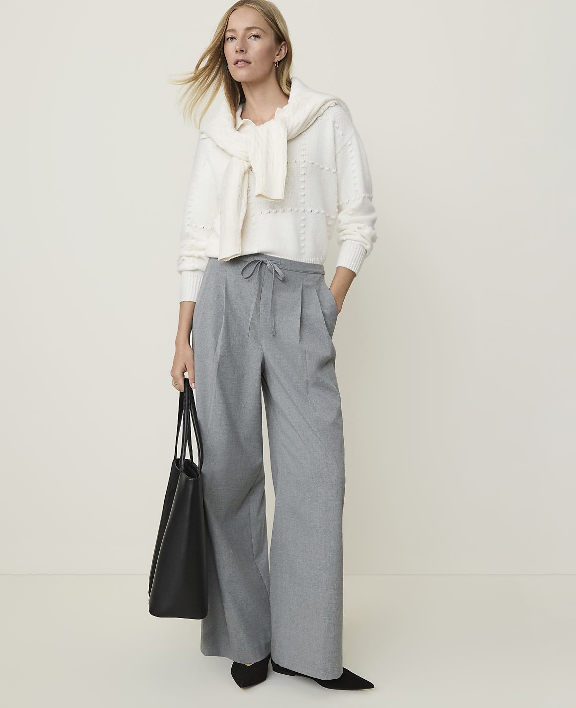 A woman in a white sweater and gray wide-leg pants stands with a black tote bag against a plain, light background.