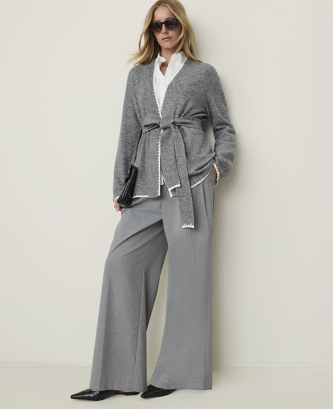 A woman in a grey belted cardigan and wide-leg pants stands against a plain, light-colored wall.