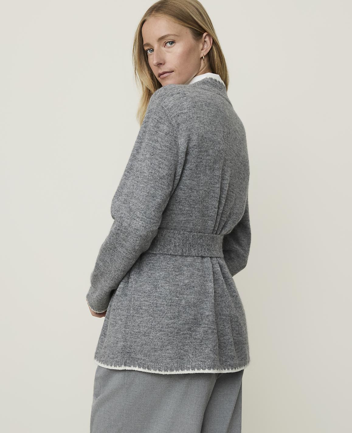 A woman in a grey belted cardigan stands against a plain, light-colored background.