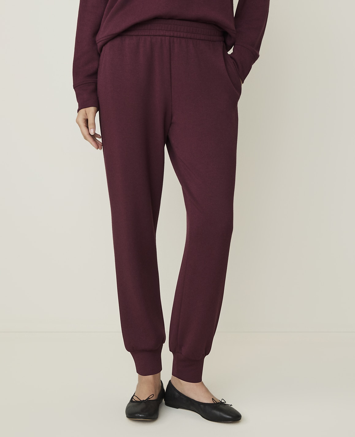 A person wearing burgundy sweatpants and black ballet flats stands against a plain, light-colored background.