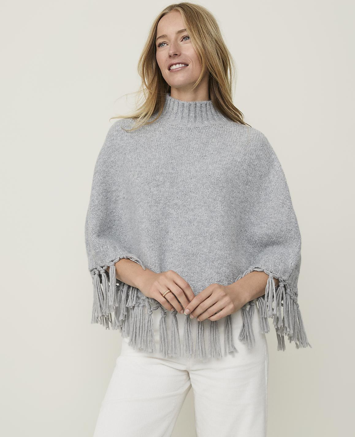 A blonde woman in a grey fringed poncho and white pants smiles against a plain, light grey background.