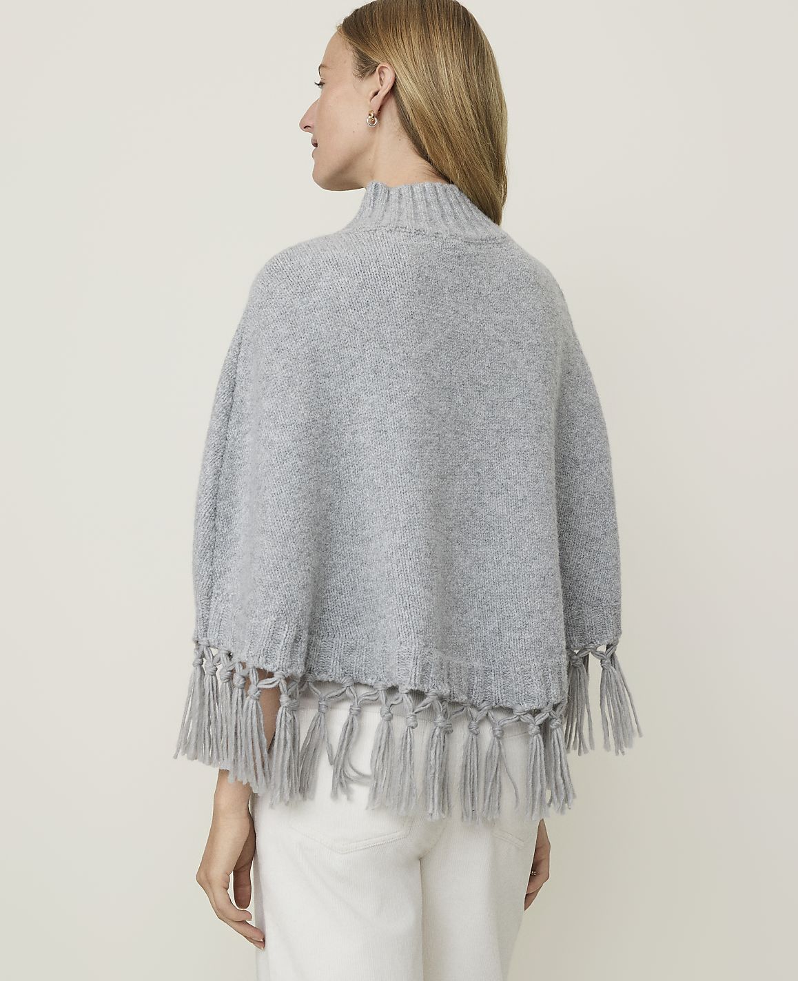 A woman in a grey poncho with fringe stands against a plain grey background.
