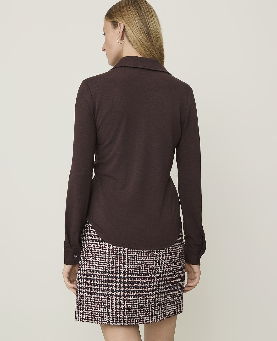 A woman in a brown collared shirt and a plaid skirt stands against a plain, light brown background.